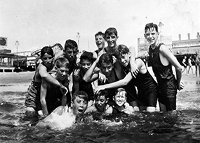 A group of 12 boys in bathing suits pose together on the beach at Coney Island.  Some boys are submerged in the water.  
